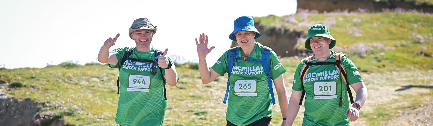 Three Mighty Hikers are walking along a pebble path. They are smiling and wearing green Macmillan branded tops. Behind them are green sea-side cliffs.