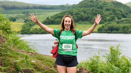 A Mighty Hiker in a celebratory pose with both arms in the air. Behind them are the lush rolling hills of the Lake District.