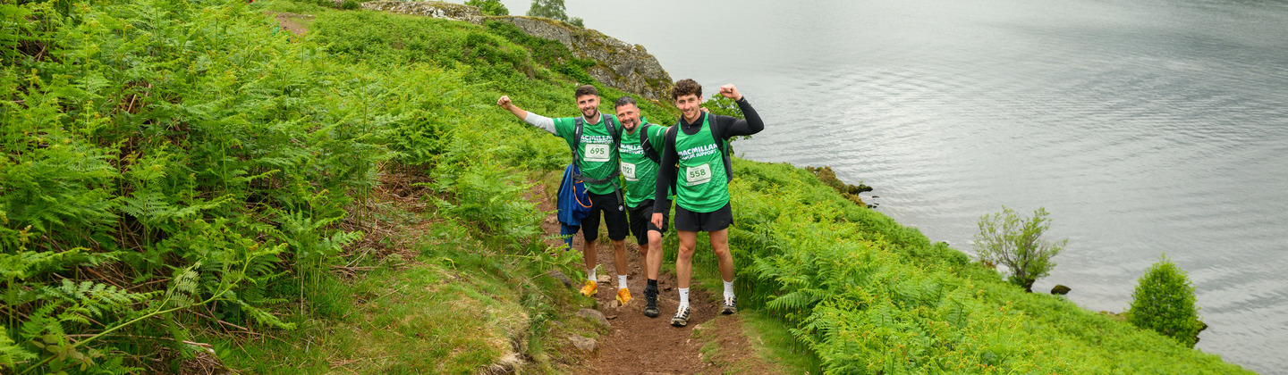 Three Mighty Hikers are stopped and posing. They are on a trail alongside a lake. They are surrounded by the lush, green rolling hills of the iconic Lake District.