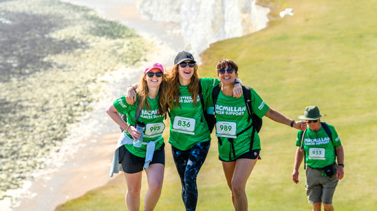 Three Mighty Hikers are standing side by side, with their arms wrapped around another. They are walking up a green hill. In the background is the iconic views of the South Coast - beach, sea side cliffs, and green hills.