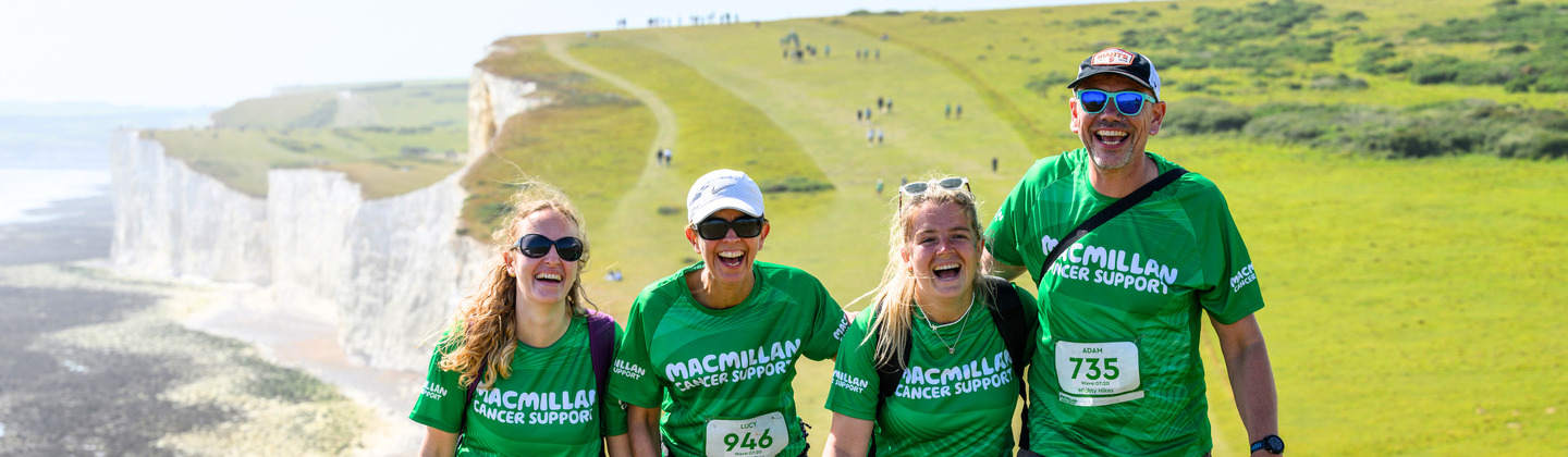 Four Mighty Hikers are trekking up a green grass hill. They are walking side by side and have bright smiles. Behind them are rolling green hills, sea side cliffs, and a beach.