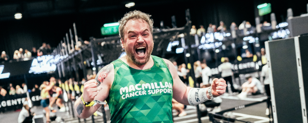 A person competing in a HYROX is cheering. They are wearing a Macmillan branded vest. Behind him are other people taking part in the HYROX.
