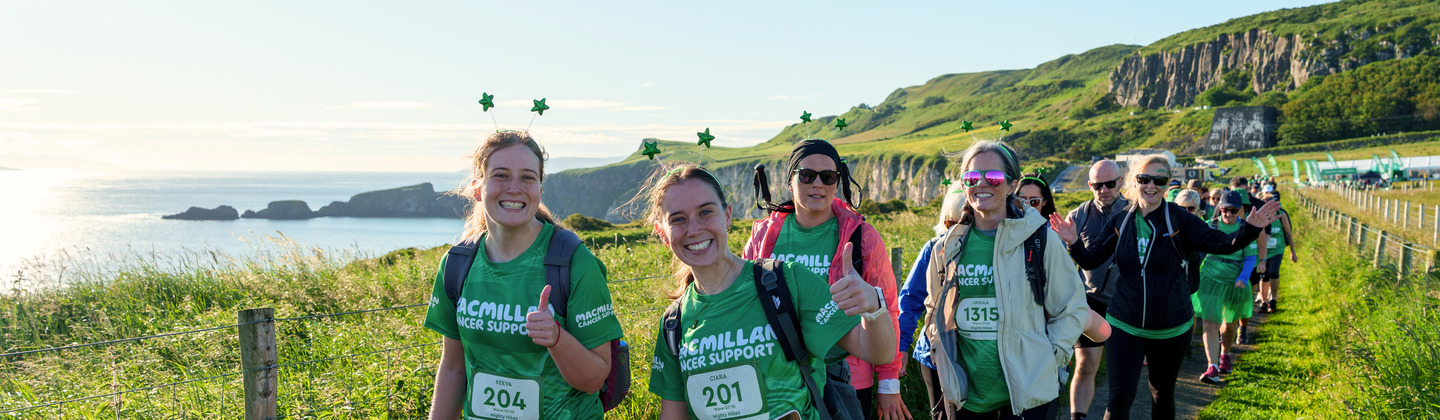 A row of Mighty Hikers are following are trekking along a dirt path. Behind them are the beautiful views of the Giants Causeway including rolling green hills and the sun shinning off the blue sea.