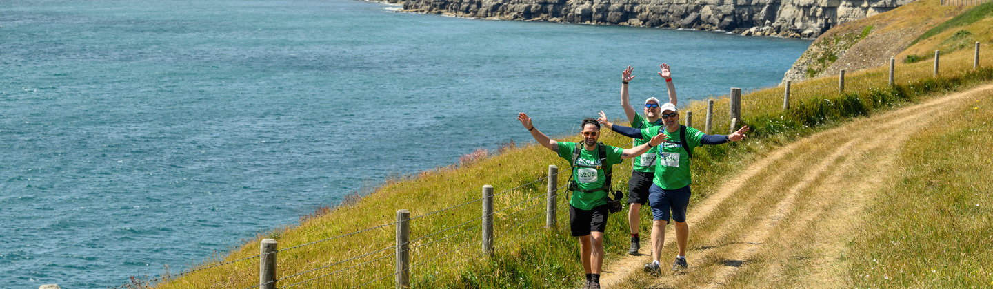 Three Mighty Hikers are trekking on a grassy trail along the sea side. They are waving their arms in the air. Behind them are the iconic views of the Jurassic Coast including rolling green hills and the clear blue sea.
