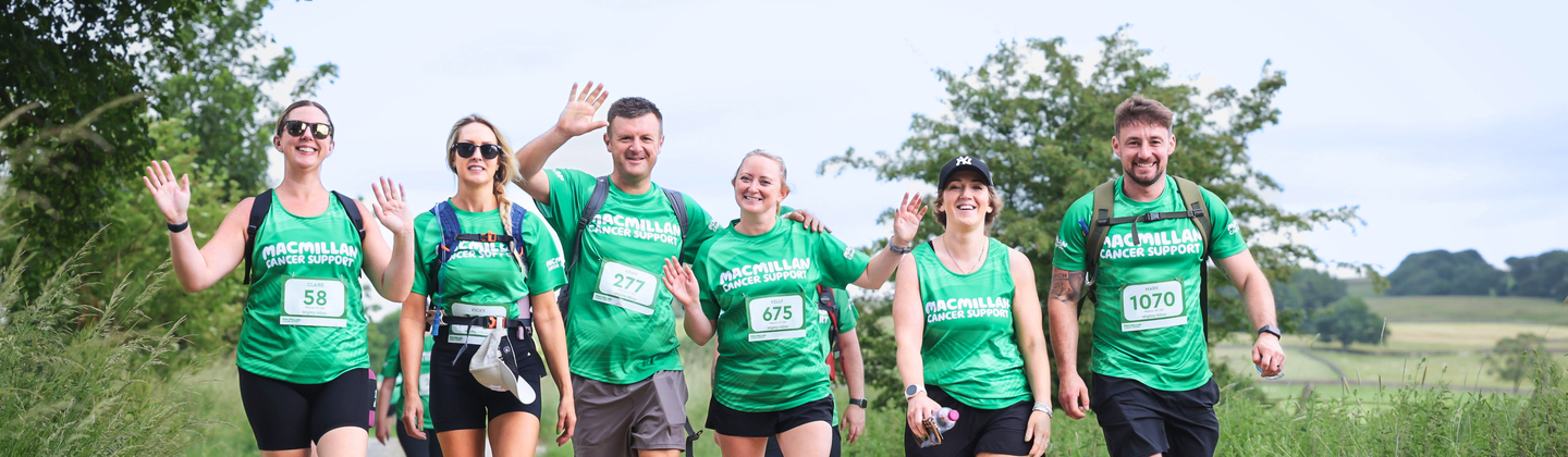 Walking side by side, a group of Mighty Hikers are trekking along a rocky path. Some of them are waving towards the camera. They are all wearing green Macmillan branded tops and other hiking gear like rucksacks.
