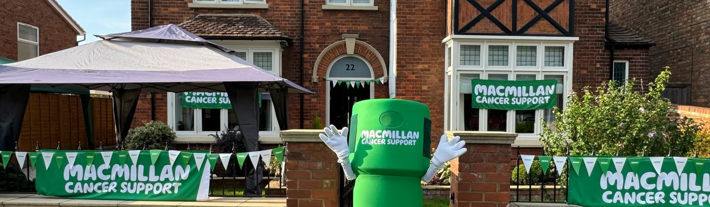 A person wearing a Macmillan branded collection tin costume is standing in front of a house decorated in green and white bunting and Macmillan Cancer Support branded banners.