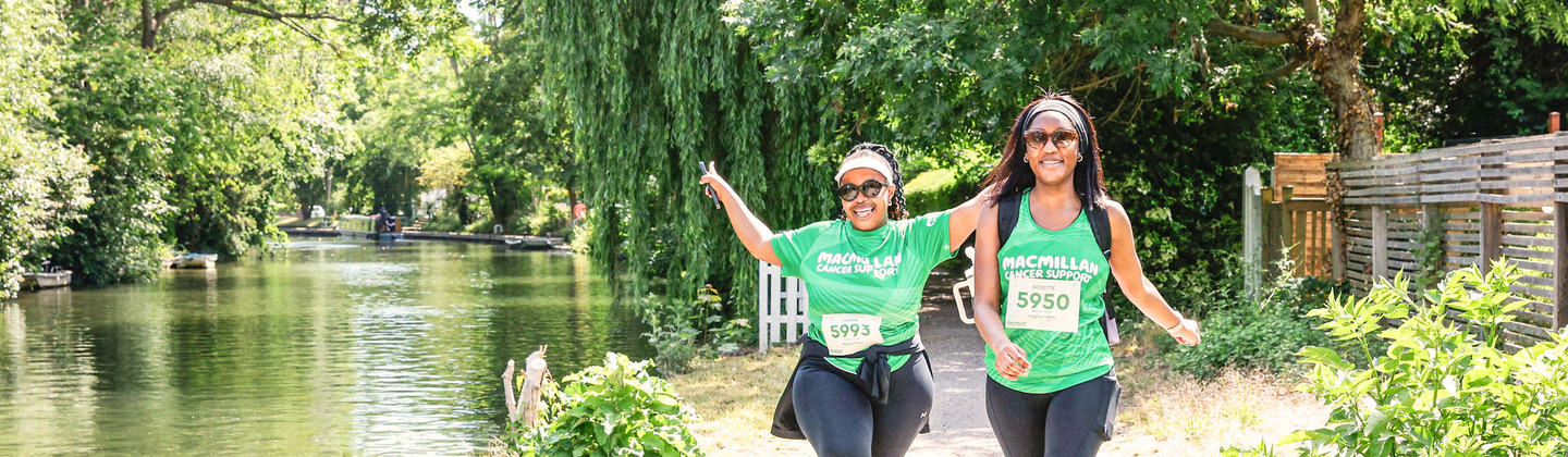 Two hikers are trekking down a path alongside a river. One of them has both arms in the air in celebration.