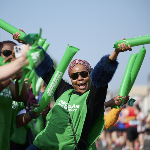 A Macmillan volunteer is cheering at the London Marathon. They have both arms in the air and are holding Macmillan branded balloons. They are wearing a long dark blue top and a Macmillan branded vest on top of it. The volunteer also has on a headscarf and sunglasses. There are other people cheering near them at the event.