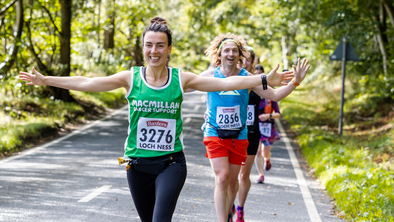 A woman with her arms outstretched running on a road and wearing a green Macmillan running vest