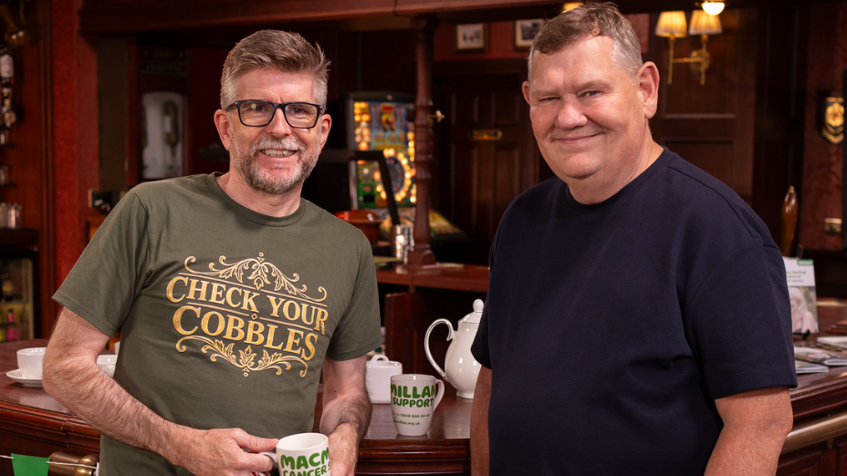 Two people inside the Rovers Return Inn with Macmillan Cancer Support mugs during the Check Your Cobbles campaign.