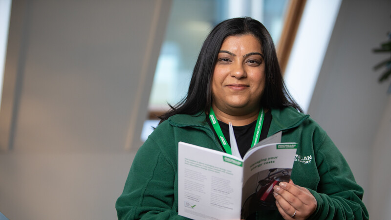 A Macmillan healthcare professional wearing a green Macmillan Cancer Support fleece and lanyard holds an information booklet in a healthcare setting. The image highlights the support, guidance and information available to people living with cancer and their loved ones.