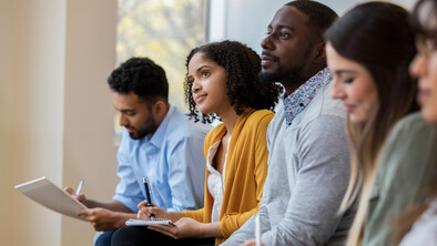 A group of people sit side by side in a bright meeting or workshop setting, taking notes and listening attentively. Several participants hold notebooks or tablets, with large windows in the background letting in natural light.