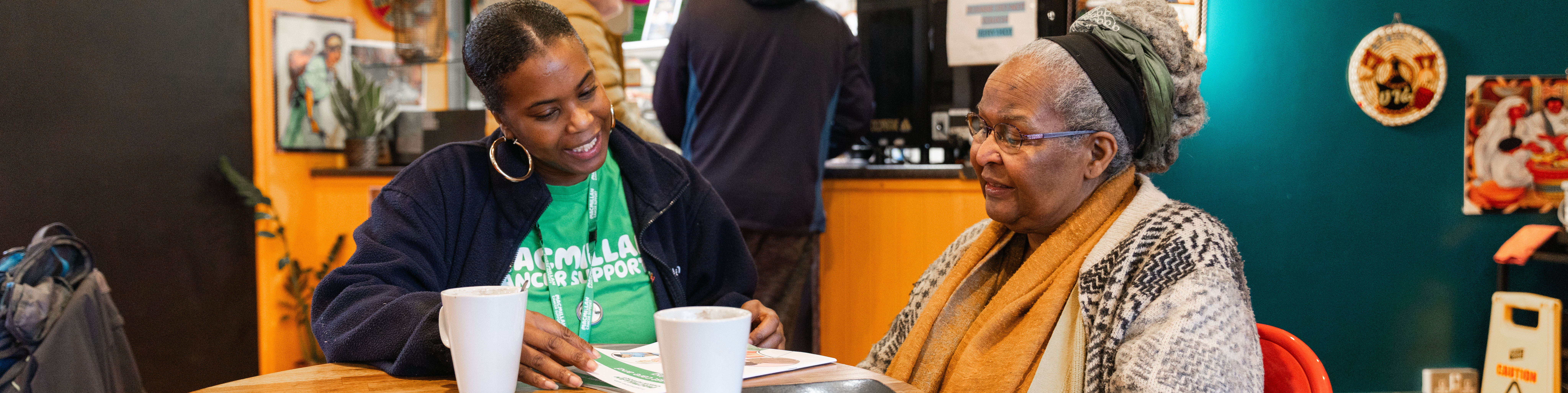 Two women sitting around a table in a cafe. One is wearing a green Macmillan Cancer Support t-shirt and lanyard.