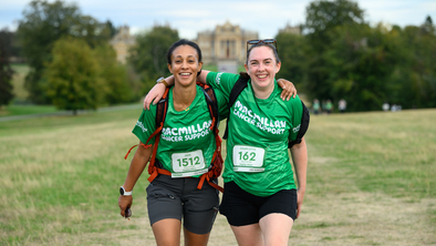 Two women with their arms around each other and smiling as they are walking, wearing their green Macmillan t-shirts.
