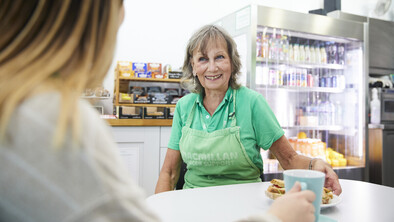 Jenny is sat at a table in the Macmillan Horizon Centre's café. She is wearing a green polo shirt and a green apron with the Macmillan logo on it. She appears to be talking to someone.