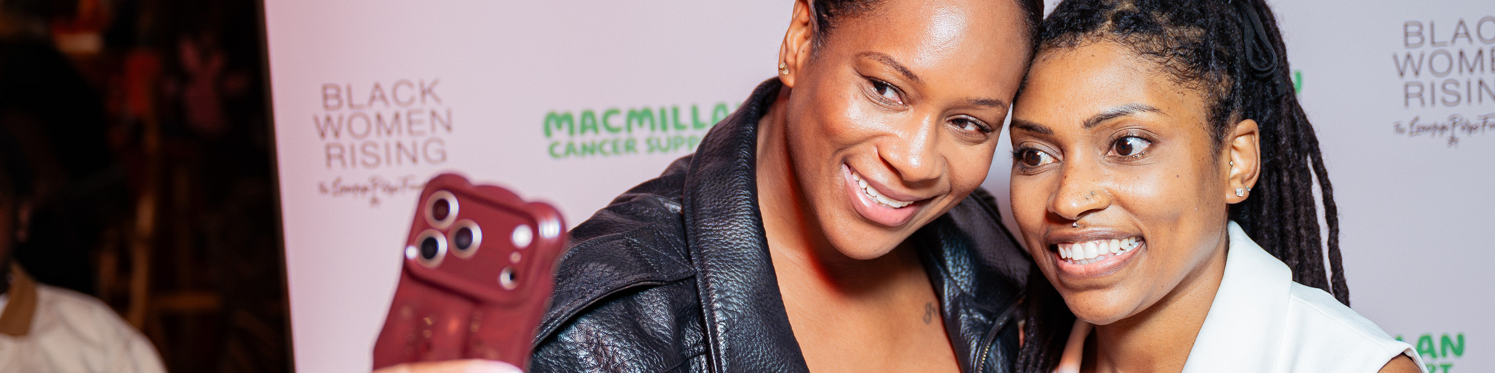 Two women pose together in front of a Black Women Rising and Macmillan Cancer Support event backdrop.
