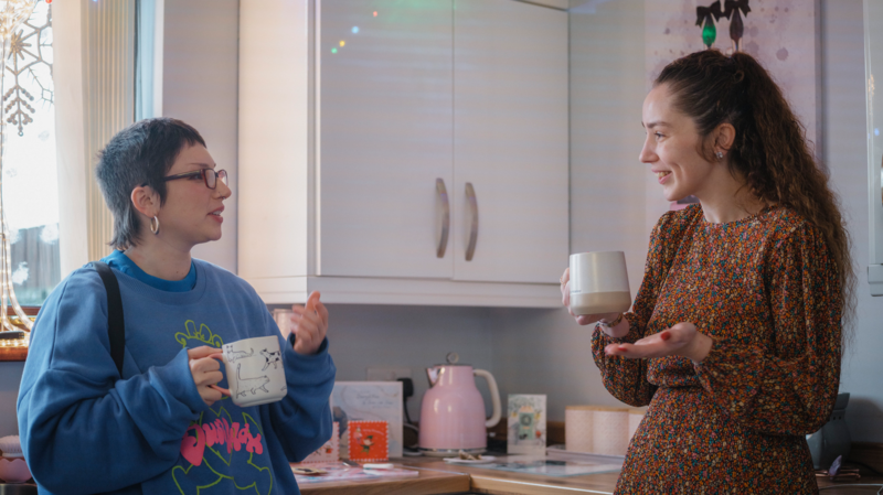 Two people stand in a kitchen holding mugs, talking casually beside a countertop decorated with lights and cards.