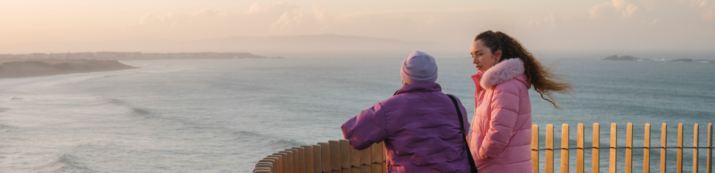 Two people in warm coats stand at a wooden viewpoint overlooking the sea, watching waves roll along a coastal shoreline at sunset.
