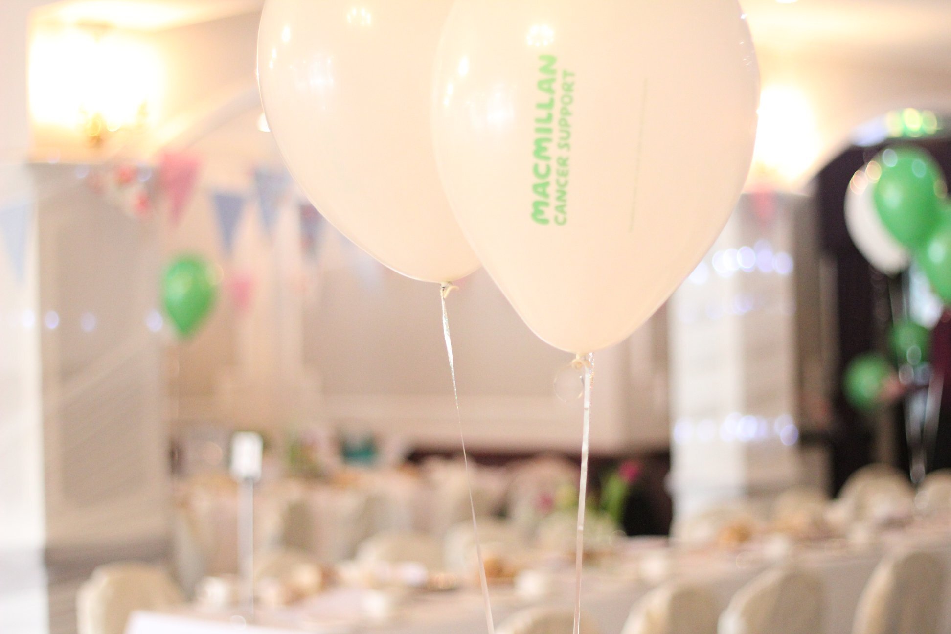 White Macmillan helium balloons are at the forefront of the photo. The rest of the image is blurred with a long table in the background set up ready for an event. 