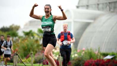 A woman wearing a Macmillan running vest running and flexing her muscles as she runs past a glass house in Kew Gardens.