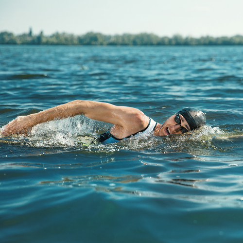 A person swimming in a lake. They are doing a free style stroke and are wearing a swimming costume, goggles and swim cap.