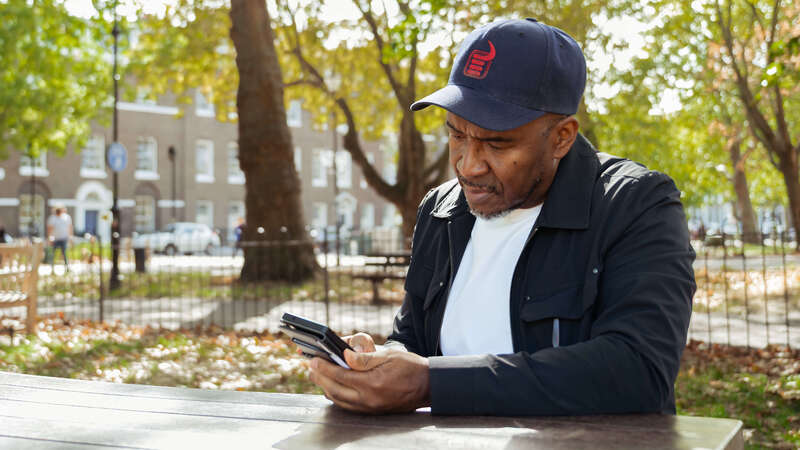 A person sits at a picnic table in a park while looking at a mobile phone, surrounded by trees and open space.