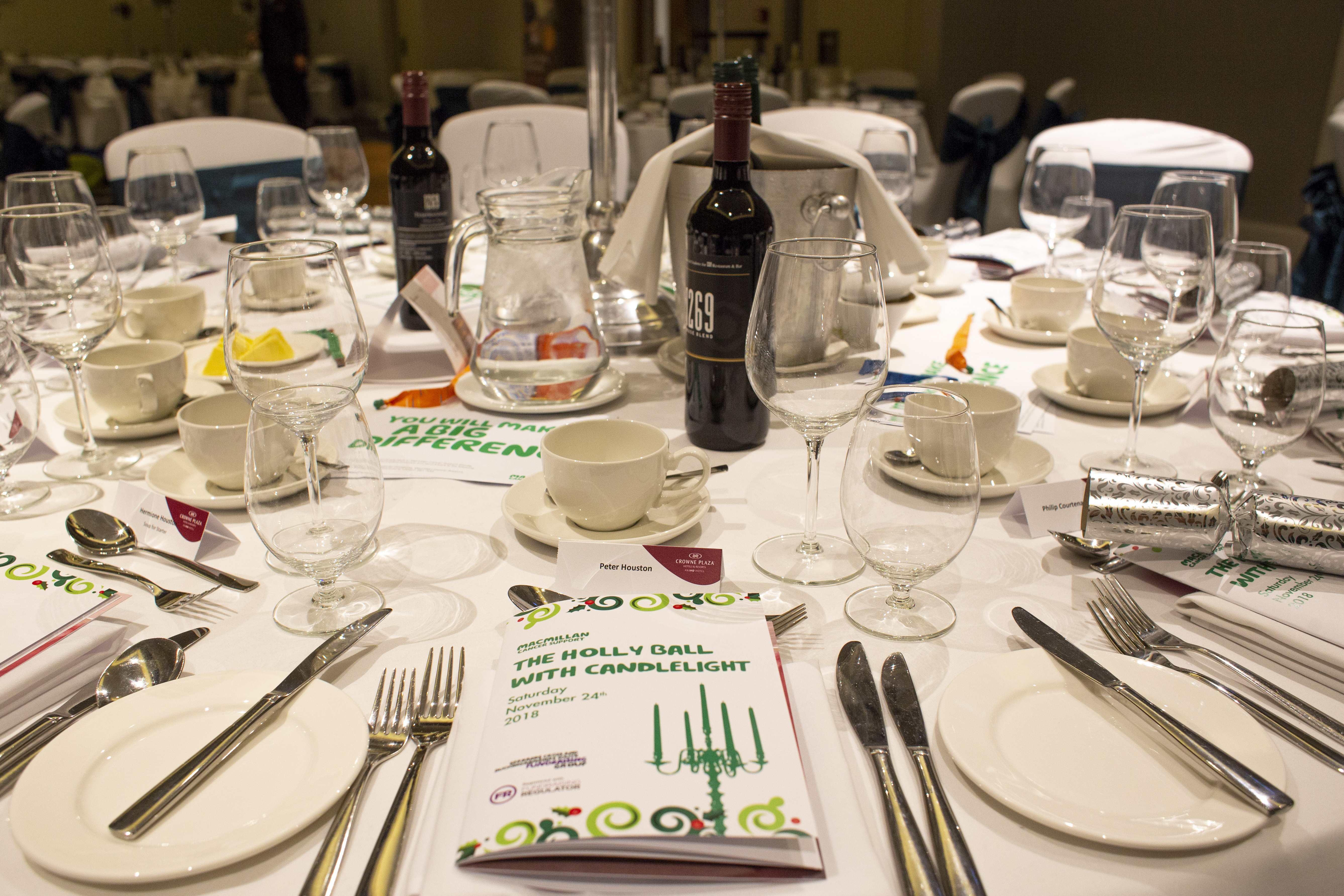 A close up image of a table with cutlery, plates and glassware ready for an event. There is an event booklet on the table that reads 'The holly ball with candlelight'. 