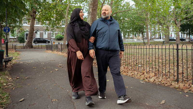 Two people walk side by side through a park, talking together in a calm outdoor setting surrounded by trees.