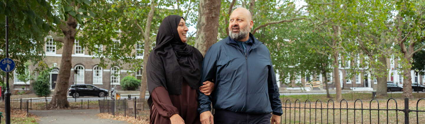 Two people walk side by side through a park, talking together in a calm outdoor setting surrounded by trees.