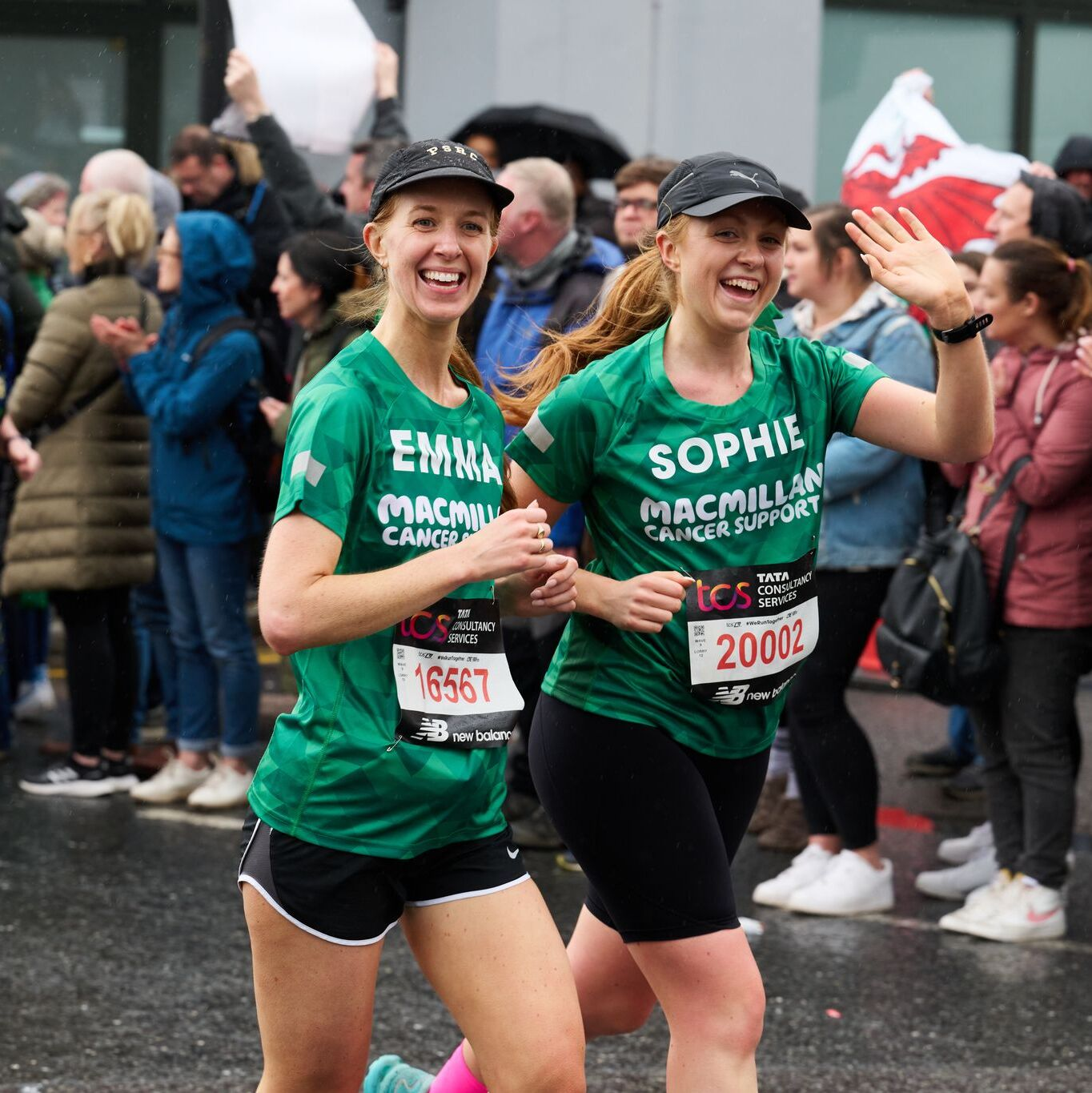 Two people are running together at an event. They are both wearing Macmillan branded green tops, caps and running shorts. People behind them are cheering on participants.