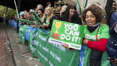 A group of volunteers are standing behind a barrier at a fundraising event. They are cheering on participants. One volunteer is holding a sign that says you can do it.