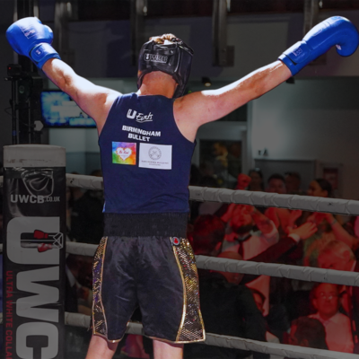 A person wearing boxing gloves has their arms in the air and is facing away from the camera. They are in a boxing ring and people are cheering for them. They are also wearing a blue top, dark shorts, and a helmet to protect their head when boxing.