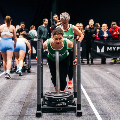 Two women wearing Macmillan vests at a HYROX event. One is do a sled push
