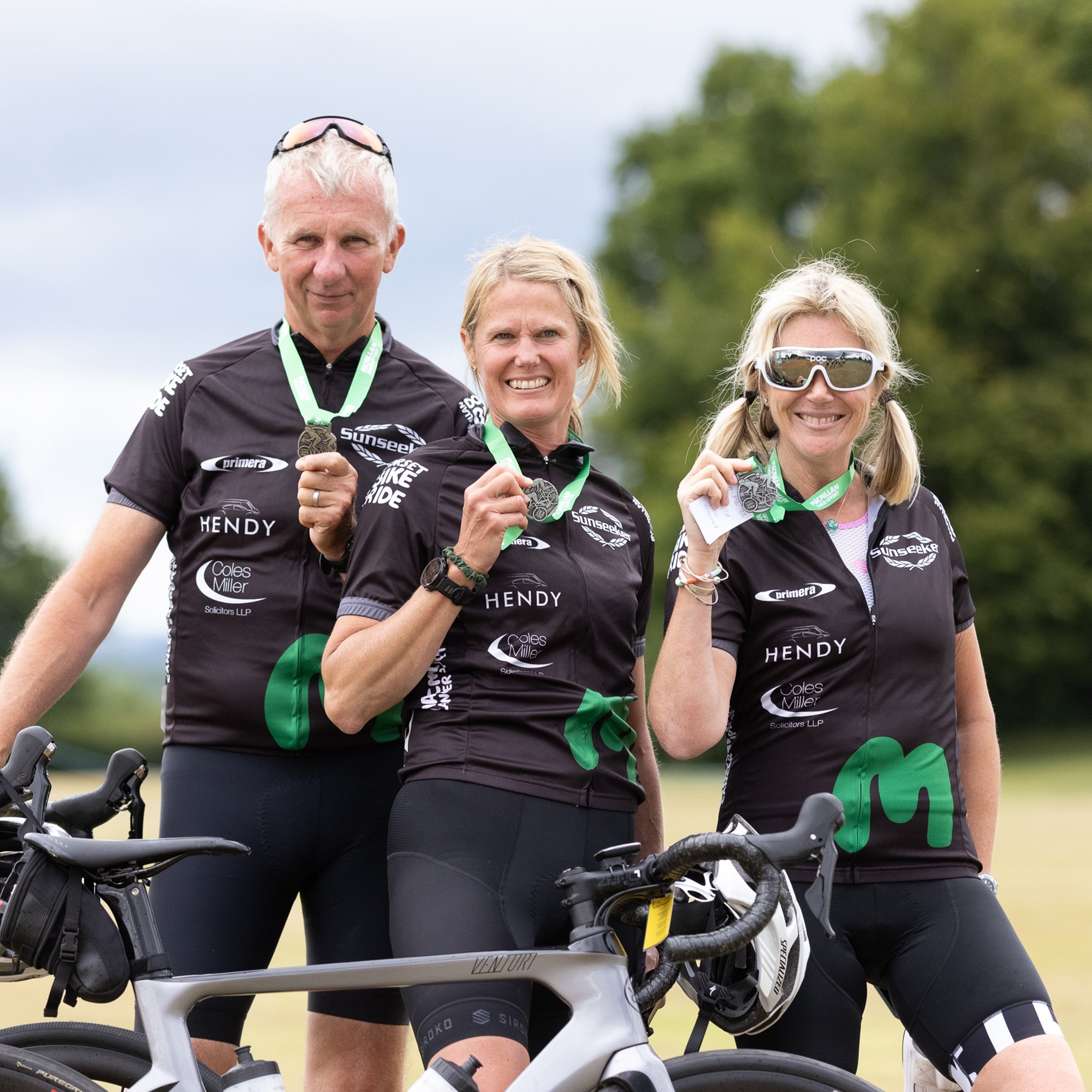 Two women and a man standing with their bikes, holding up medals and wearing their Macmillan cycling tops
