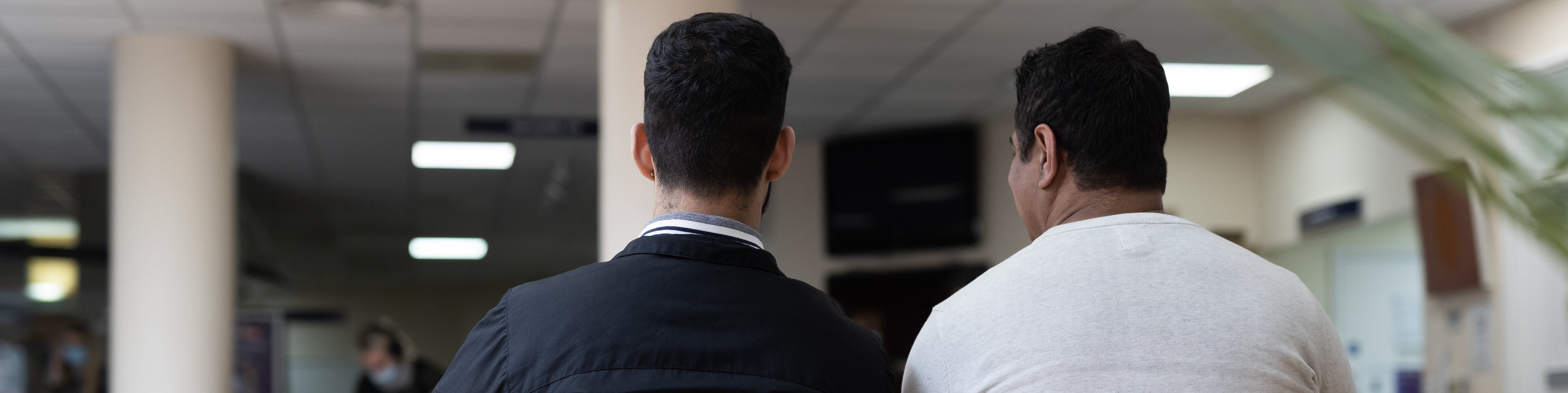 Two men with their backs to the camera. They are in a hospital setting sitting on orange seats in a waiting room.