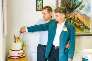 There are 2 well dressed men on their wedding day embracing each other as they cut their wedding cake. 