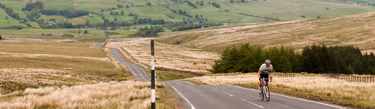 A man cycles up an empty road surrounded by an expansive countryside landscape.