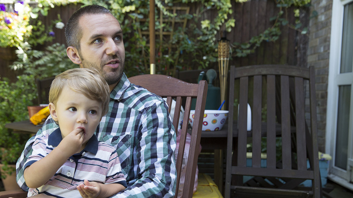 Luke is sat in the garden with his son on his lap.