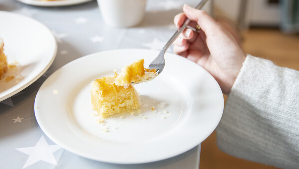 Slice of lemon cake on a white plate. A hand holds a fork with a piece of cake on it ready to be eaten.