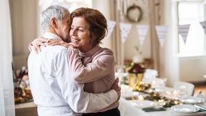 An older man and a woman hug. Behind them, a table laid for a party can be seen and bunting hangs overhead.