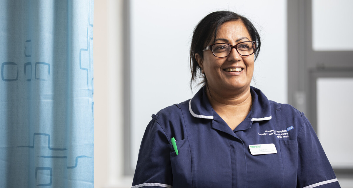 A female Macmillan Nurse wearing a blue uniform stands next to a hospital cubicle curtain