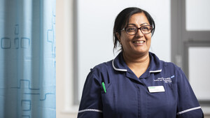 A female Macmillan Nurse wearing a blue uniform stands next to a hospital cubicle curtain