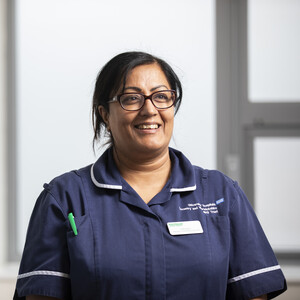 A female Macmillan Nurse wearing a blue uniform stands next to a hospital cubicle curtain