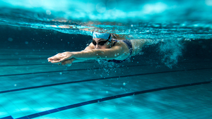 A woman swims with her hands out in front having just dived into the pool. She wears a swim cap and goggles.