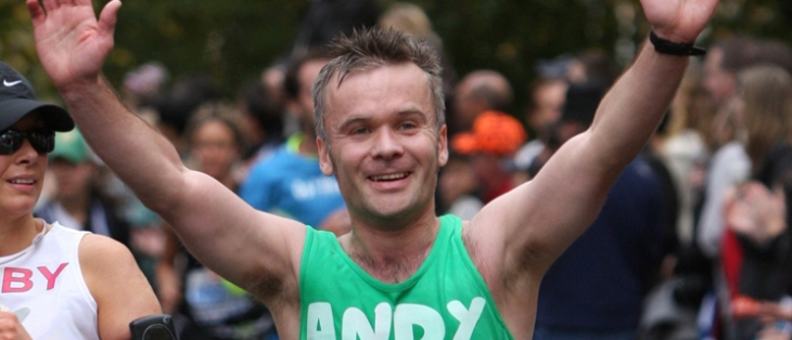 Man wearing a green Macmillan running vest with Andy on the front smiling and with both hands in the air.