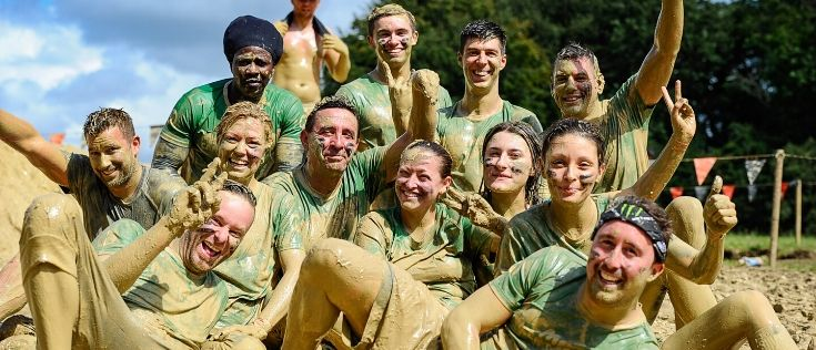 Group of 12 people covered from head to toe in mud smiling at the camera wearing green Macmillan t-shirts.