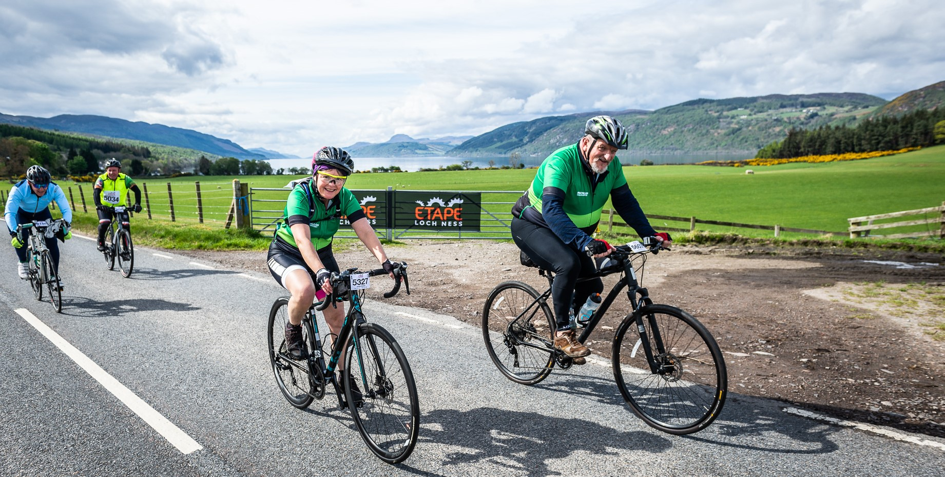 Two people wearing green cycling tops riding along a road and smiling at the camera with a Loch in the background.