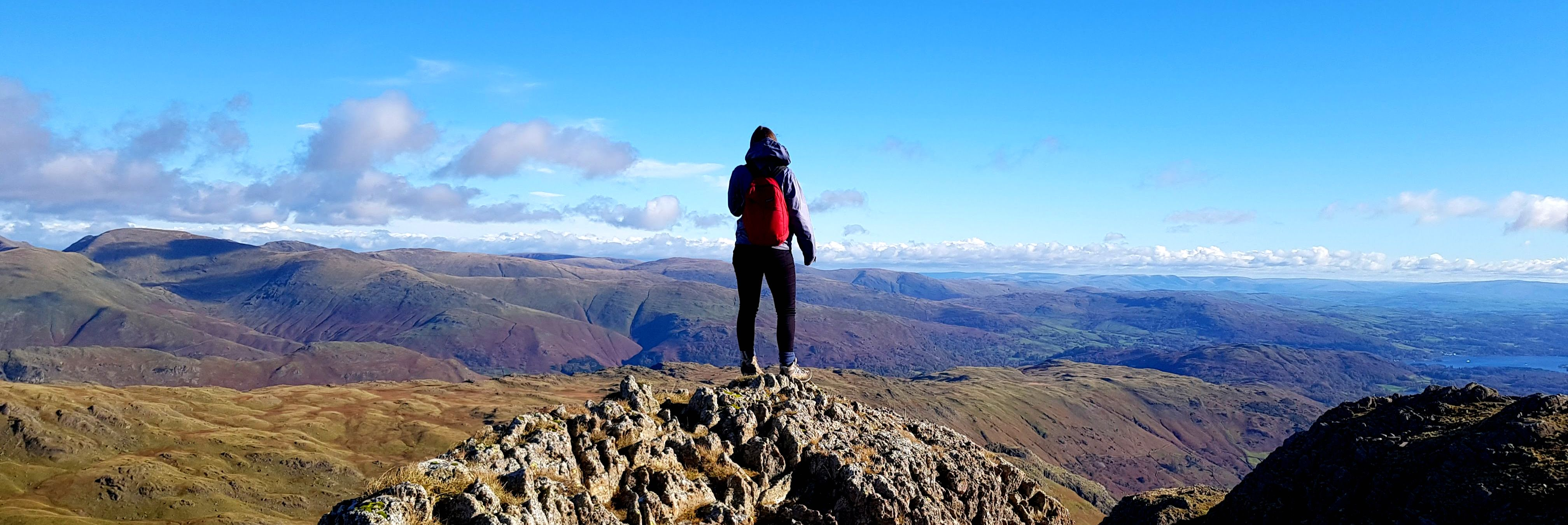 Someone with a red backpack standing on top of a large hill looking out across a hilly landscape