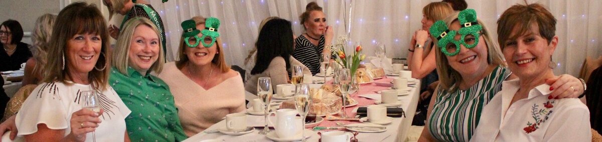 A group of ladies sat around a table smiling. 2 of the ladies are wearing novelty green glasses. 