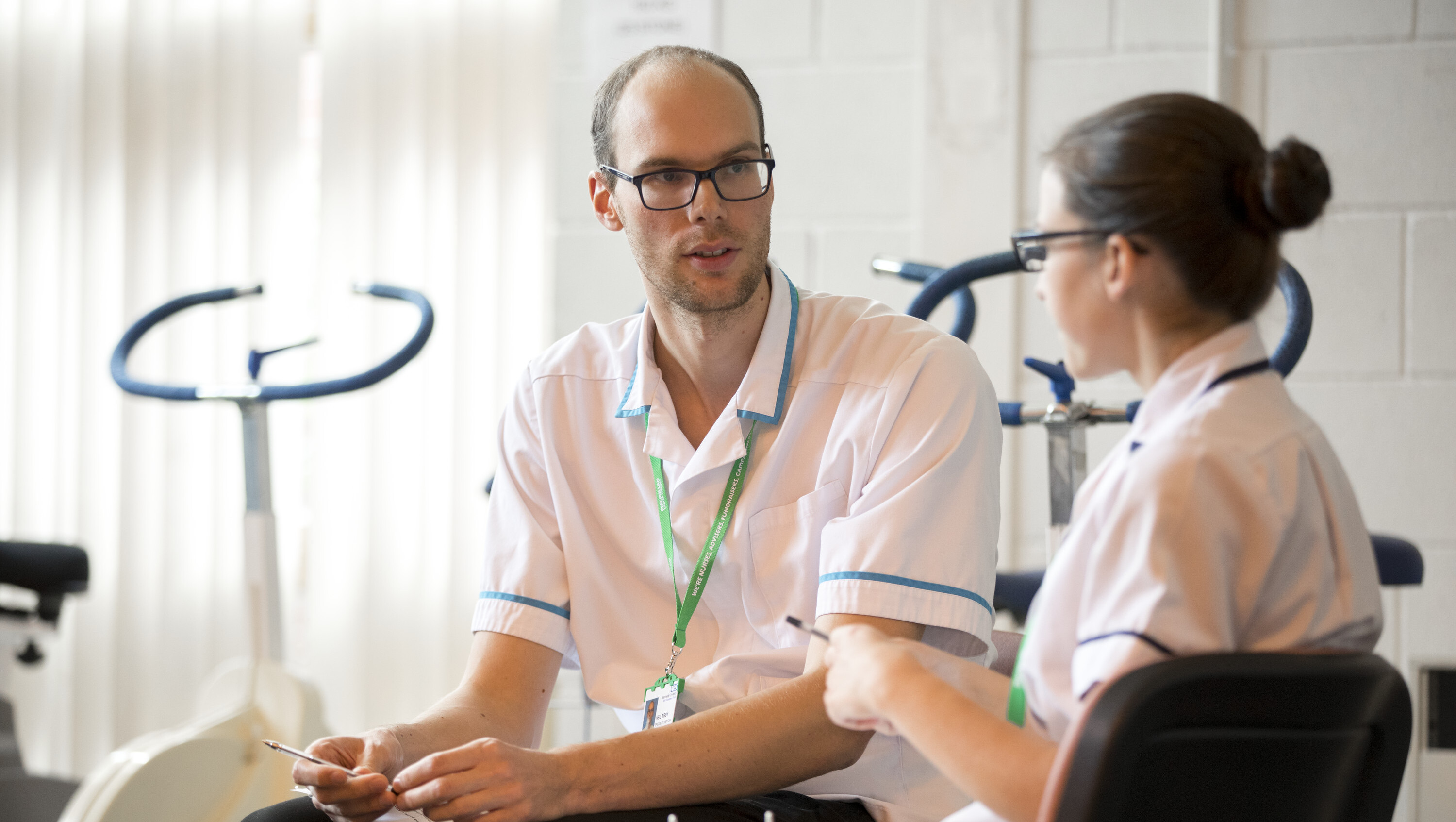 Two Allied Health Professionals wearing white tunics and green Macmillan lanyards sitting and talking to eachother. There are exercise bikes in the background.